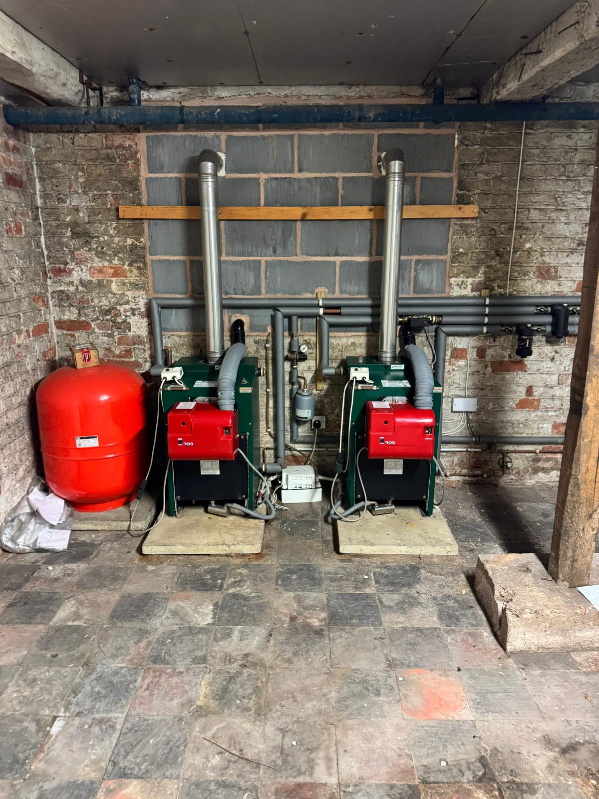 Two floor-standing oil boilers installed side by side in a brick cellar, with flue pipes, insulated pipework and a red expansion vessel